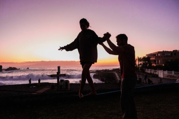 Couple healping each other to learn how to balance on slackline in Tenerife park – rental for group activities